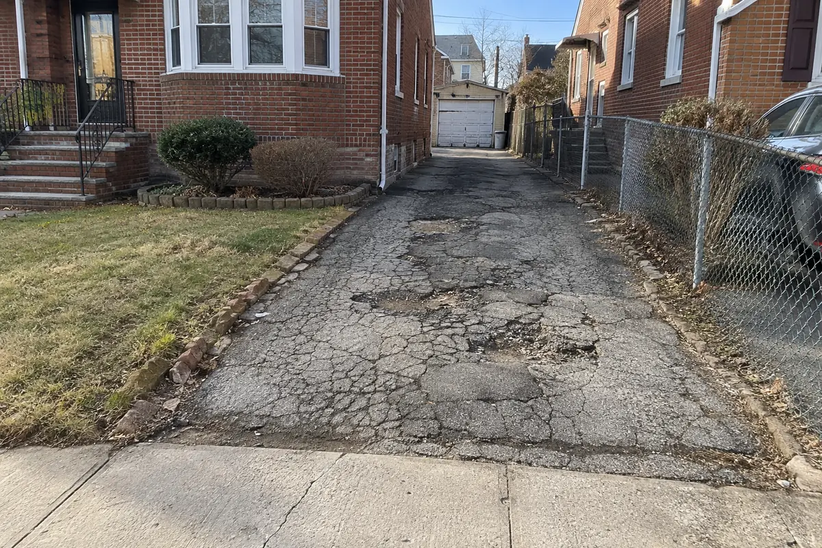 Residential asphalt driveway with extensive cracking, deteriorated pavement, and uneven surface areas leading to a detached garage.