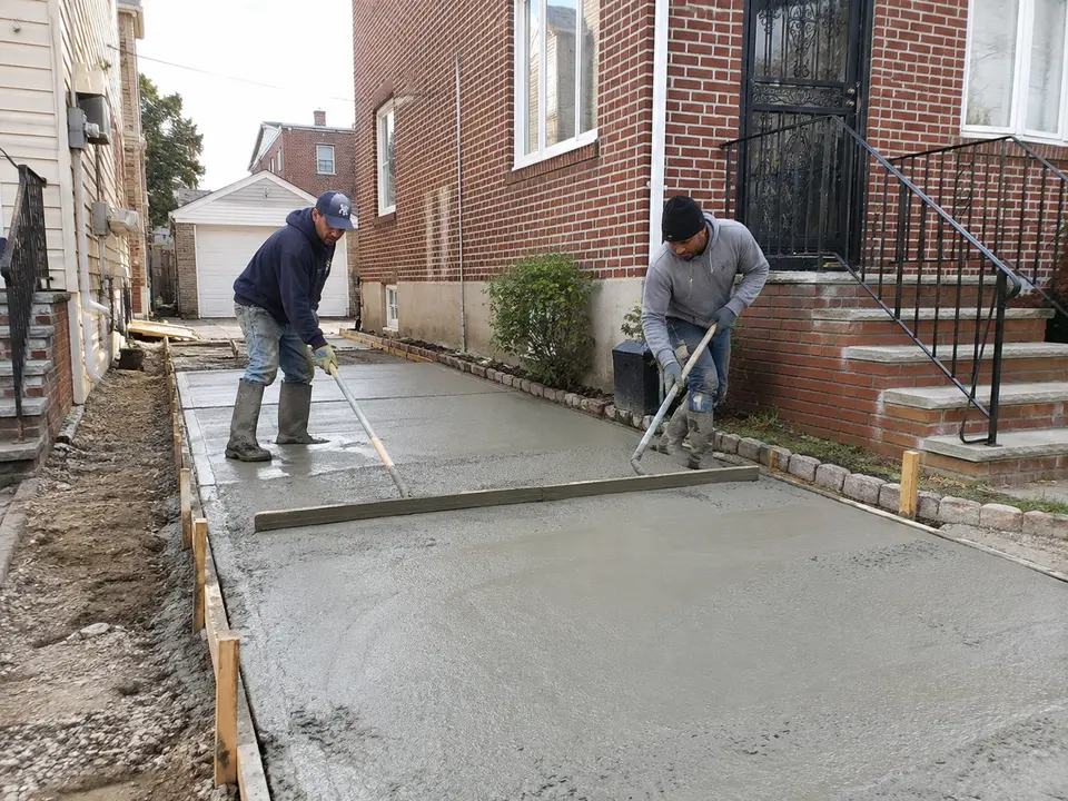Workers spreading and leveling freshly placed concrete for a residential driveway beside a brick home.