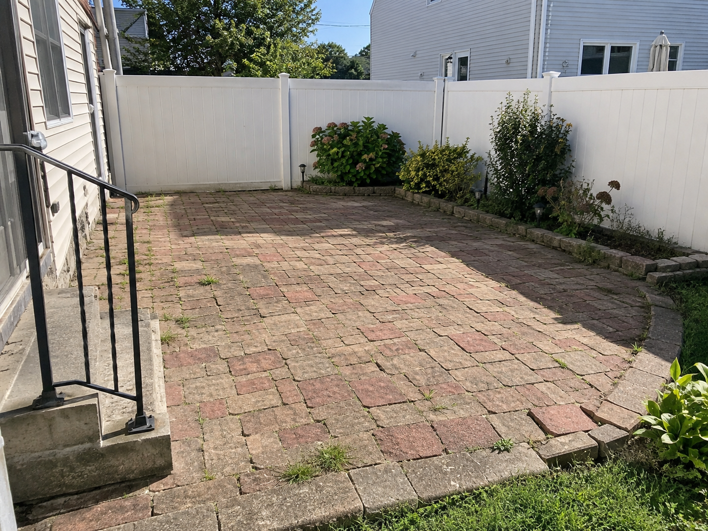 Aging brick-paver patio with uneven and weathered paving stones beside a white residential privacy fence.
