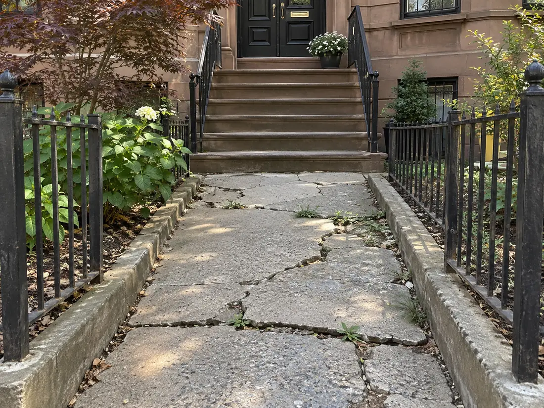 Cracked and uneven concrete entrance walkway outside a Brooklyn brownstone with damaged pavement and residential steps.