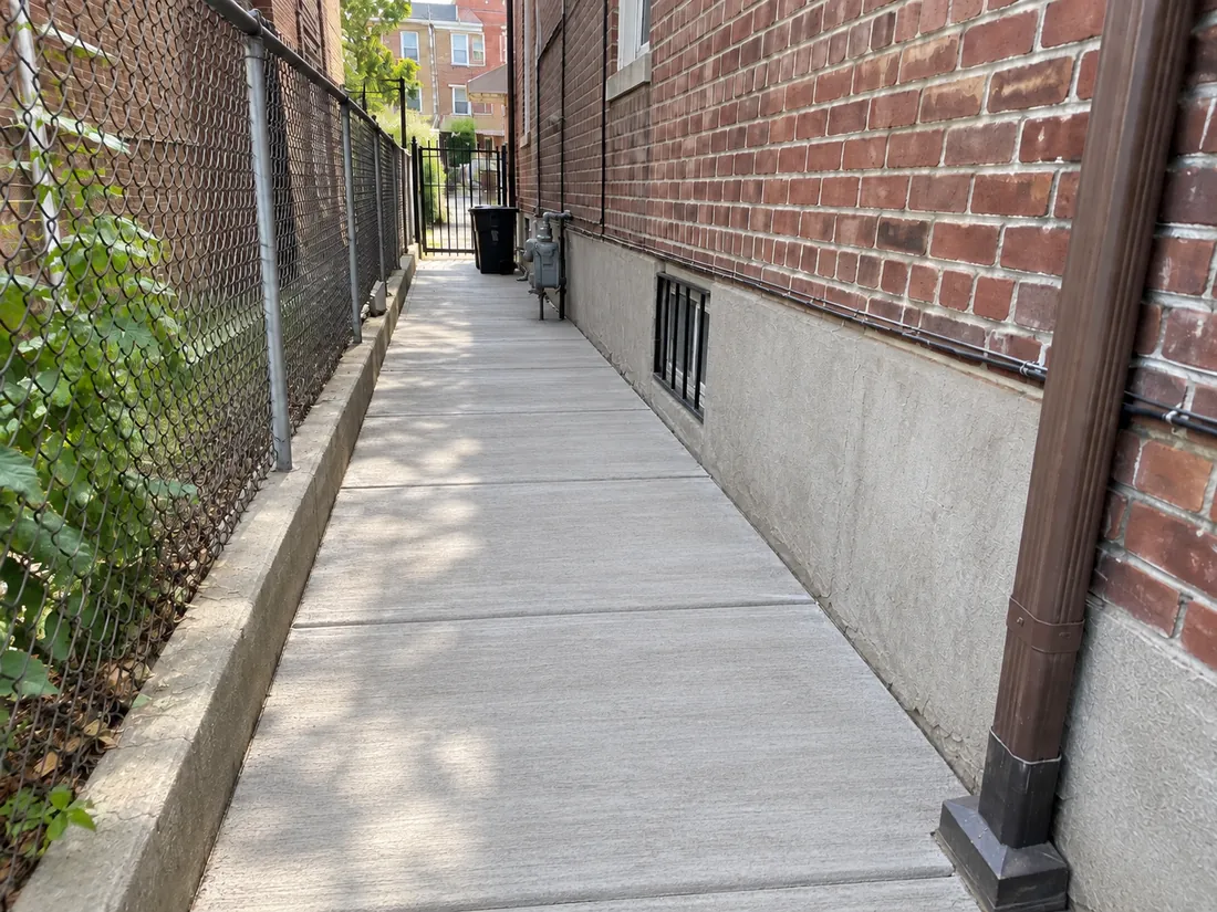 Newly installed concrete side-access walkway with a broom-finished surface and clean control joints beside an NYC residential building.