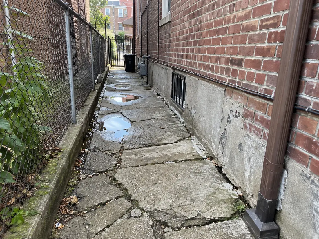 Damaged concrete side-access walkway with cracked pavement and standing water between residential buildings in NYC.